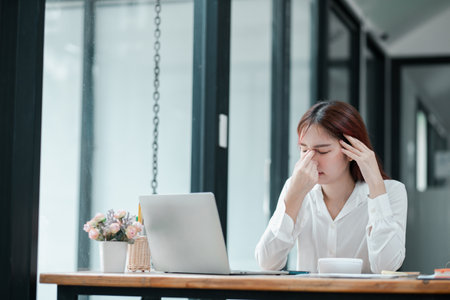 A young professional woman in a modern office setting, showing signs of stress while working on her laptop.の写真素材