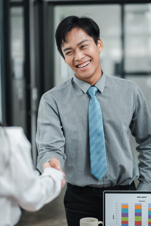 A cheerful businessman in a gray shirt and blue tie shaking hands with a colleague in a modern...