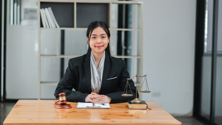 A young female lawyer in a professional office setting, seated at a desk with legal scales and a gavel, exuding confidence and professionalism.の写真素材