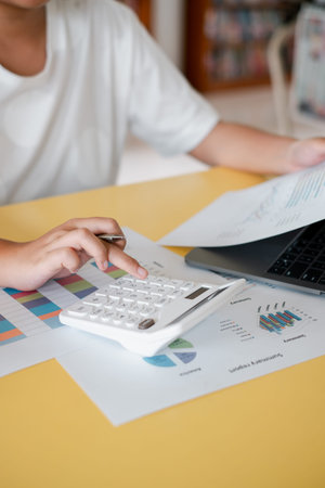 Close-up of a person using a calculator and laptop for financial analysis, surrounded by graphs and...