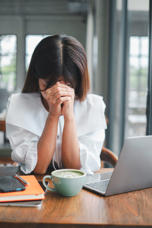 A woman sits at a cafe table with a laptop and coffee, appearing stressed and overwhelmed, with her...