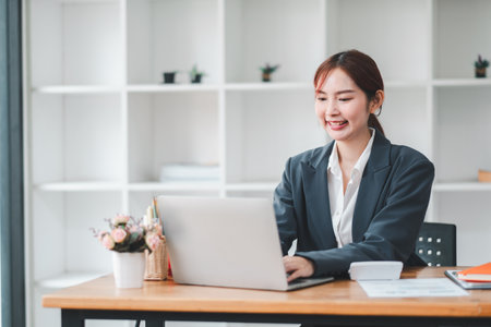 Smiling young woman in business attire working on a laptop at a desk in a contemporary office setting.の写真素材