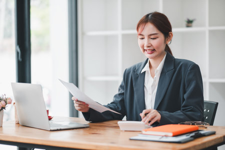 A young professional woman in a business suit working at her desk with a laptop and documents in a modern office environment.の写真素材