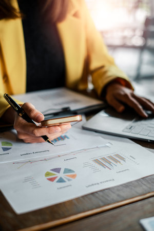 Close-up of a business professional reviewing financial charts and graphs using a smartphone and laptop in a modern office environment.の写真素材