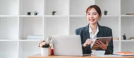 Smiling young professional woman using a tablet while working in a modern office setting with a laptop and organized shelves in the background.の写真素材