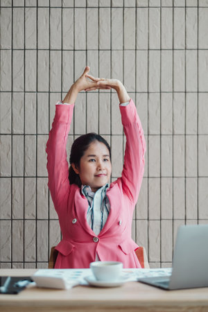Professional woman in pink blazer stretching at her desk in a modern office setting, with a laptop and coffee cup.の写真素材