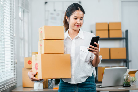 A young woman using a smartphone to manage online business orders while holding and packing boxes in a home office setting.の写真素材
