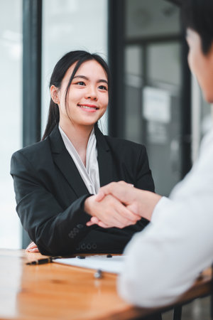 Professional businesswoman in a black suit shaking hands with a colleague in a modern office environment, symbolizing successful business interaction.の写真素材