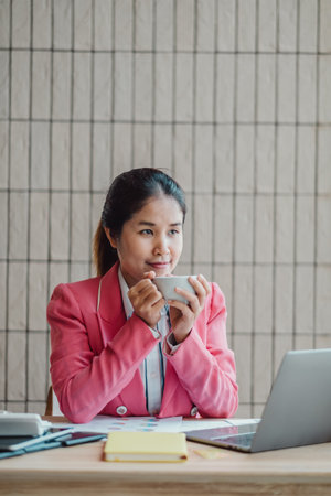 Professional woman in a pink blazer enjoying a coffee break while working on her laptop at a modern office desk.の写真素材