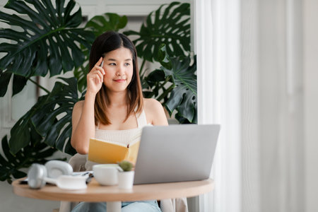 A young woman sits at a table with a laptop and notebook, working from home in a bright, modern room with large green plants.の写真素材