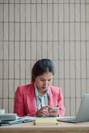 Professional woman in a pink blazer focused on her smartphone at a modern office desk with a laptop and documents.の写真素材