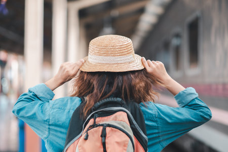 A person with a backpack and straw hat stands at a train station platform, ready for travel and...