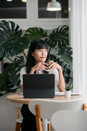 A young woman sits at a cafe table with a tablet, enjoying a coffee break surrounded by lush green...