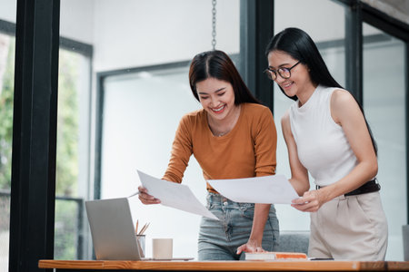 Two young women smiling and working together on a project in a bright, modern office space.の写真素材