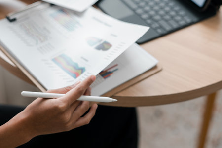 Close-up of a person reviewing financial charts with a stylus and tablet in a contemporary workspace.の写真素材