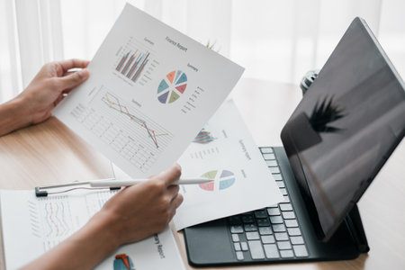 Close-up of hands reviewing financial charts and graphs on paper with a laptop on a desk in a bright office.の写真素材