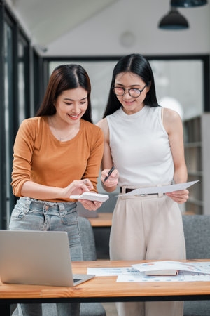 Two young women working together in a bright office, discussing documents and using a laptop.の写真素材