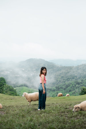 A woman stands in a green pasture with sheep, surrounded by misty mountains, creating a serene and...