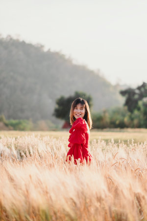 A woman in a red dress smiles while standing in a field of tall grass, with trees and hills in the background.の写真素材