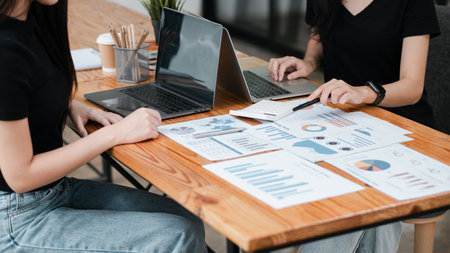 Two professionals reviewing financial documents and graphs on laptops, collaborating in a modern office environment.の写真素材
