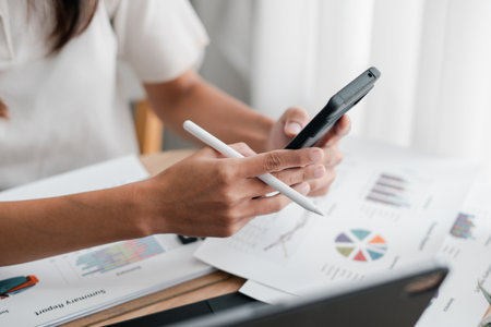 Close-up of hands using a smartphone and stylus to review financial charts and graphs on paper in a bright office.の写真素材