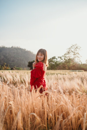A woman in a vibrant red dress stands amidst a golden wheat field, with a scenic mountain view in the background.の写真素材