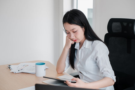 Young woman concentrating on work at a desk with a tablet and documents, showcasing a professional...