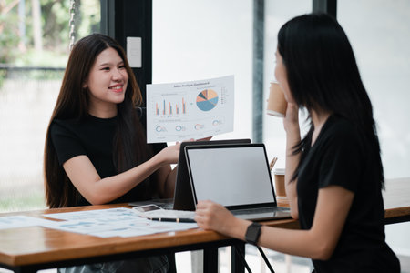 Two women engaged in a business meeting, analyzing charts and data on laptops in a contemporary office environment.の写真素材
