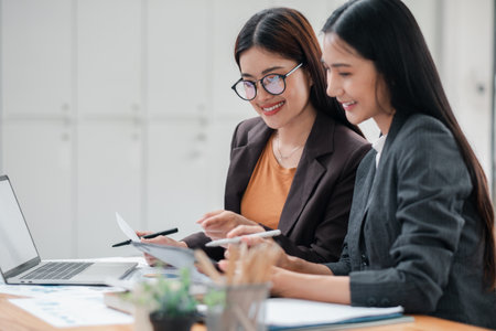 Two businesswomen working together at a desk, reviewing documents and using a laptop in a bright, modern office.の写真素材