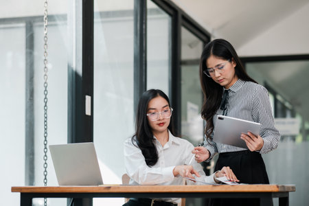Two businesswomen working together in a contemporary office setting, using a laptop and tablet for collaboration.の写真素材