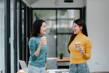 Two women in casual attire chatting and holding coffee cups in a bright, modern office environment.の写真素材