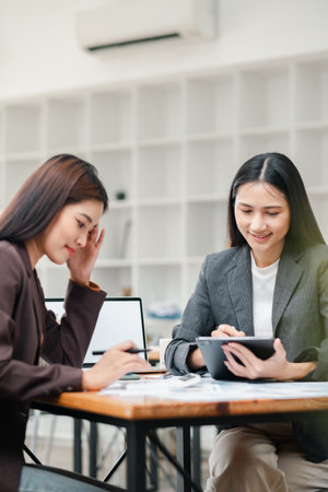 Two women in a modern office setting discussing work with laptops and documents, showcasing teamwork and collaboration.の写真素材