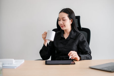 Professional woman in black attire sitting at a desk, holding a coffee mug, with a tablet and laptop...