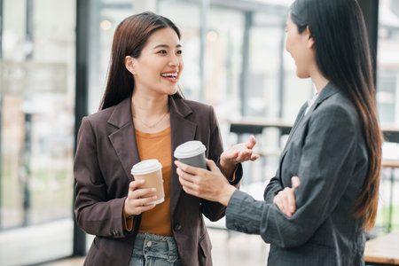 Two professional women chatting and enjoying coffee in a bright, modern office environment.の写真素材