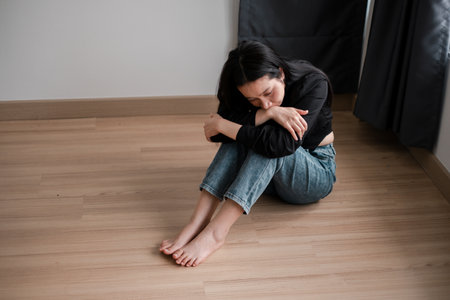 A young woman in casual clothing sits on a wooden floor, deep in thought, illuminated by natural light.の写真素材