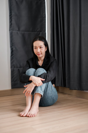 A young woman in casual attire sits thoughtfully on a wooden floor, framed by dark curtains.の写真素材