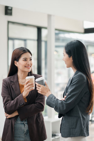 Two businesswomen in a modern office, holding coffee cups, engaging in conversation, and smiling.の写真素材