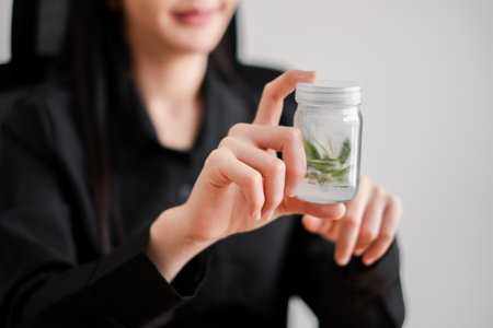 Close-up of a woman holding a jar with green leaves, symbolizing eco-friendly and sustainable living in a modern office environment.の写真素材