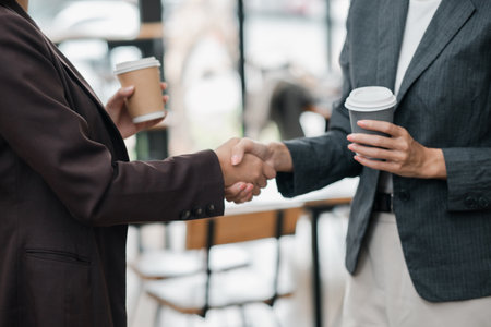 Two business professionals shaking hands in an office, holding coffee cups, symbolizing partnership and collaboration.の写真素材