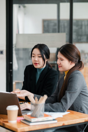 Two women in business attire working together in a contemporary office, focusing on a laptop and discussing documents.の写真素材