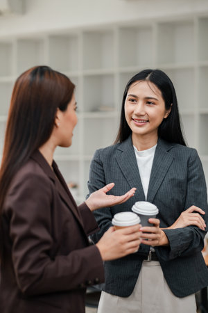 Two businesswomen in formal attire discussing work while holding coffee cups in a contemporary office environment.の写真素材
