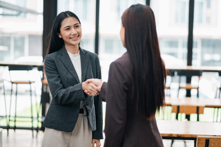 Two businesswomen in formal attire shaking hands in a bright, modern office space with large windows.の写真素材