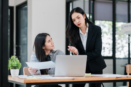 Two businesswomen in a modern office setting discussing a project, using a laptop and documents, showcasing teamwork and collaboration.の写真素材