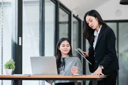 Two businesswomen in a modern office setting, discussing work with a laptop and tablet, showcasing teamwork and collaboration.の写真素材