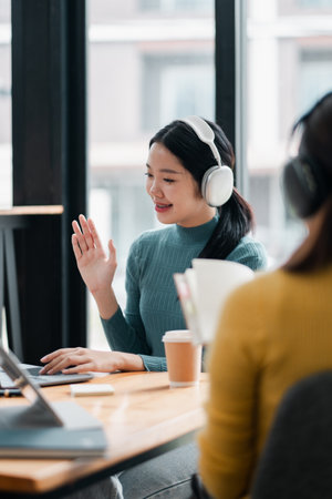 Smiling woman wearing headphones, participating in an online meeting at a contemporary office with a laptop and coffee.の写真素材