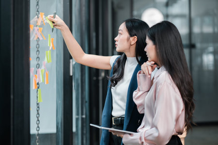 Two businesswomen brainstorming ideas on a glass wall with sticky notes in a contemporary office...