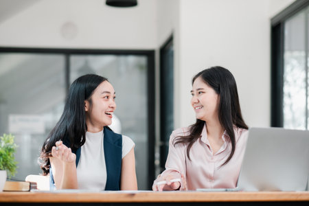 Two women smiling and talking in a bright, modern office setting with a laptop on the table.の写真素材