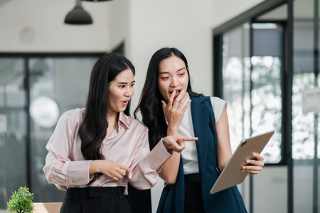 Two professional women in a modern office, engaged and excited while looking at a tablet, showcasing teamwork and innovation.の写真素材