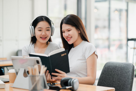 Two young women sharing a book in a cafe, surrounded by modern tech and cozy ambiance.の写真素材