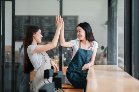 Two young women wearing aprons high-five in a modern cafe, symbolizing teamwork and success in a bright, cheerful setting.の写真素材